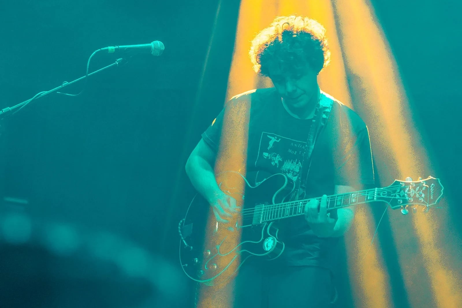 Gitarrist mit halbakustischer Gitarre im türkis-gelben Lichtstrahl auf der Bühne.