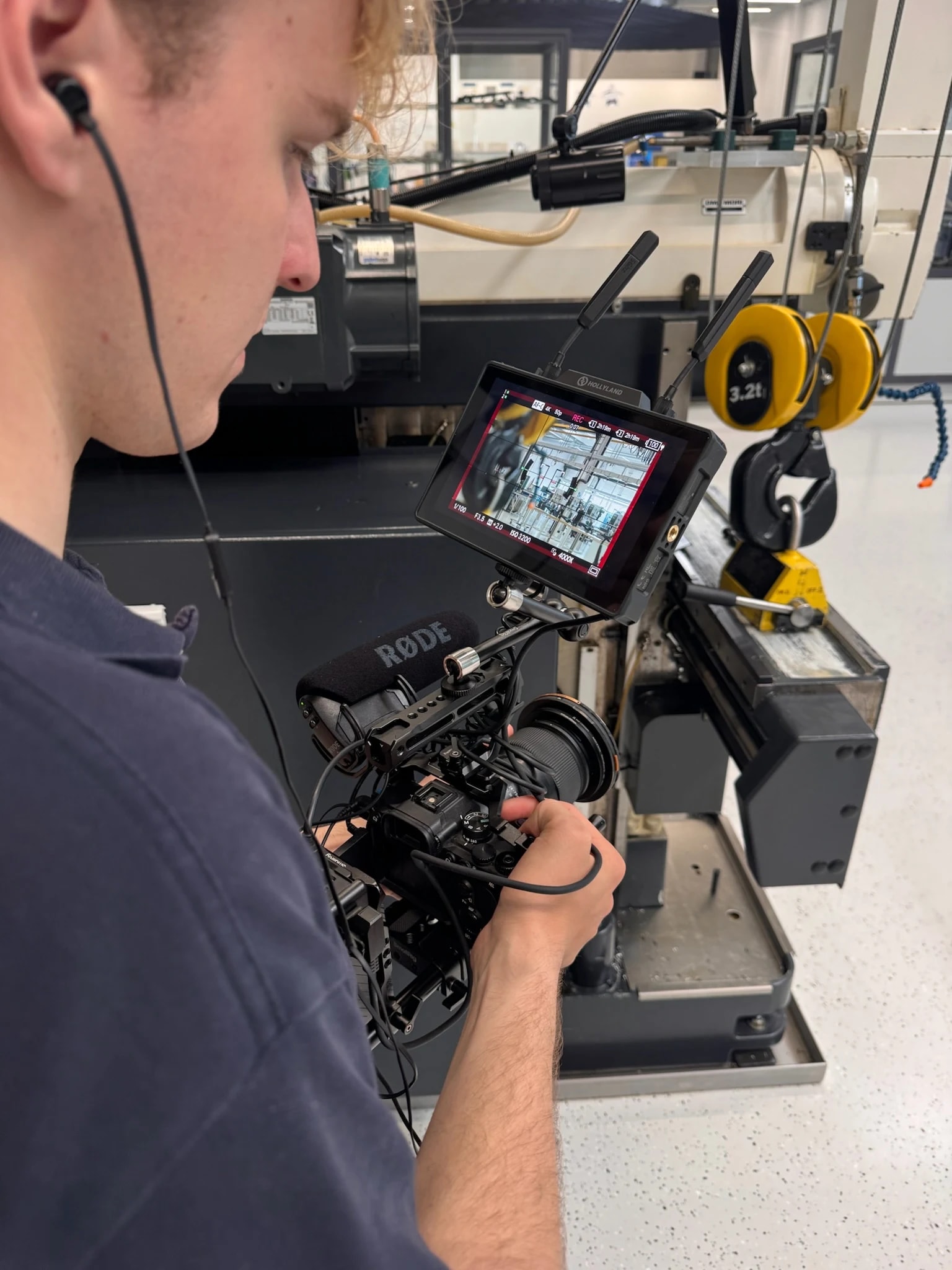 Kameramann mit In-Ear-Kopfhörern filmt mit einer Kamera samt Monitor und Richtmikrofon in der Werkshalle; daneben stehen eine Maschine und ein gelber Kranhaken.