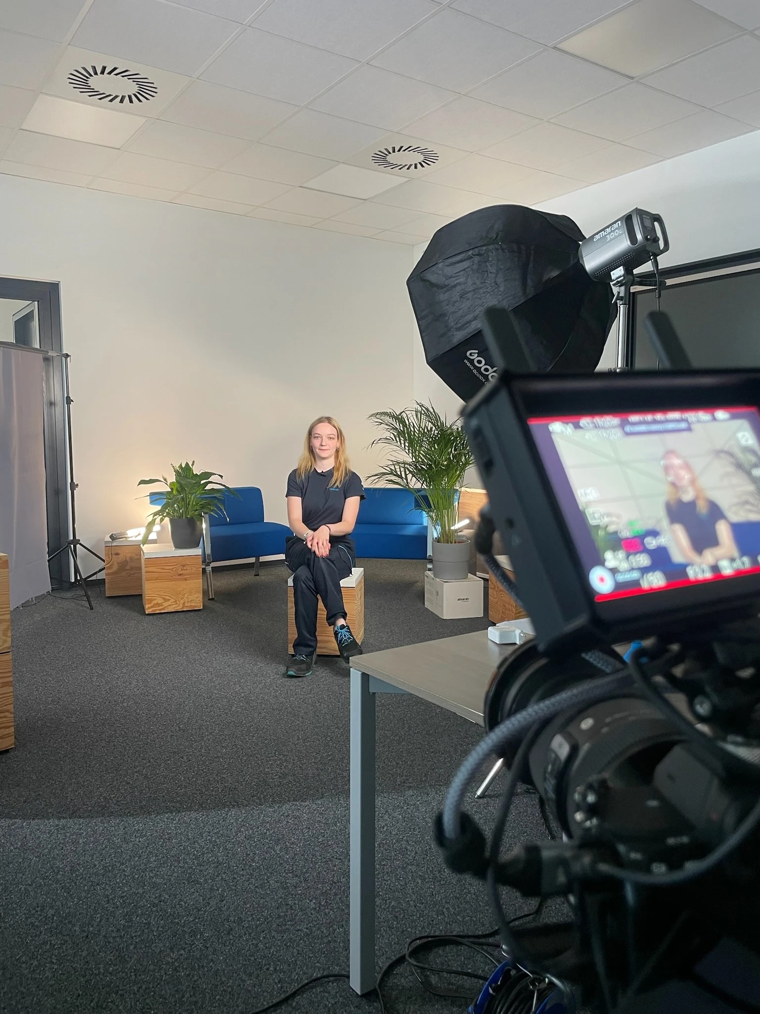 Interview-Setup mit Kamera und Softbox: Eine Lehrling sitzt auf einem Holzhocker vor blauen Sofas und Pflanzen, im Vordergrund zeigt der Kameramonitor ihr Bild.