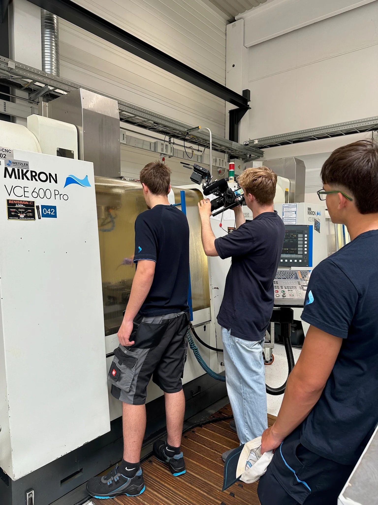 Kameramann filmt mit Schulterkamera einen Lehrling, der in das Sichtfenster einer CNC-Fräsmaschine blickt; ein weiterer Lehrling sieht im Vordergrund zu.