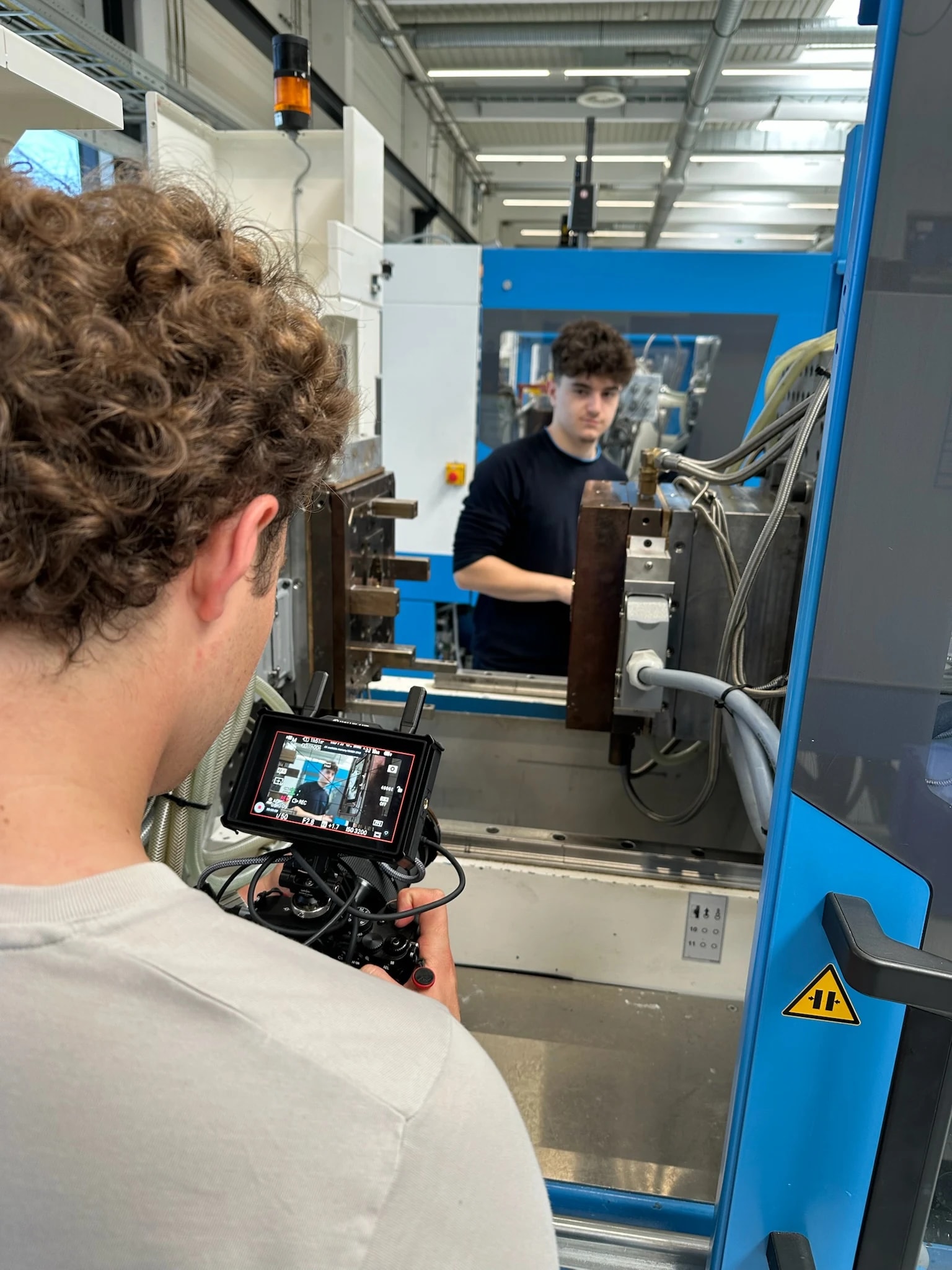 Kameramann filmt über die Schulter einen Lehrling an einer blauen Spritzgussmaschine mit geöffnetem Werkzeug; auf dem Kameramonitor ist das Bild des Lehrlings zu sehen.