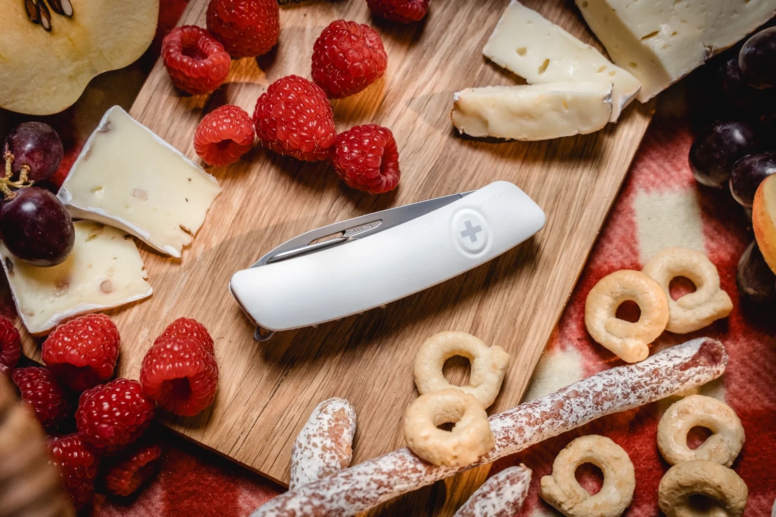 Geschlossenes SWIZA Taschenmesser mit weißem Griff auf dem Holzbrett, umgeben von Käse, Himbeeren und Trauben.