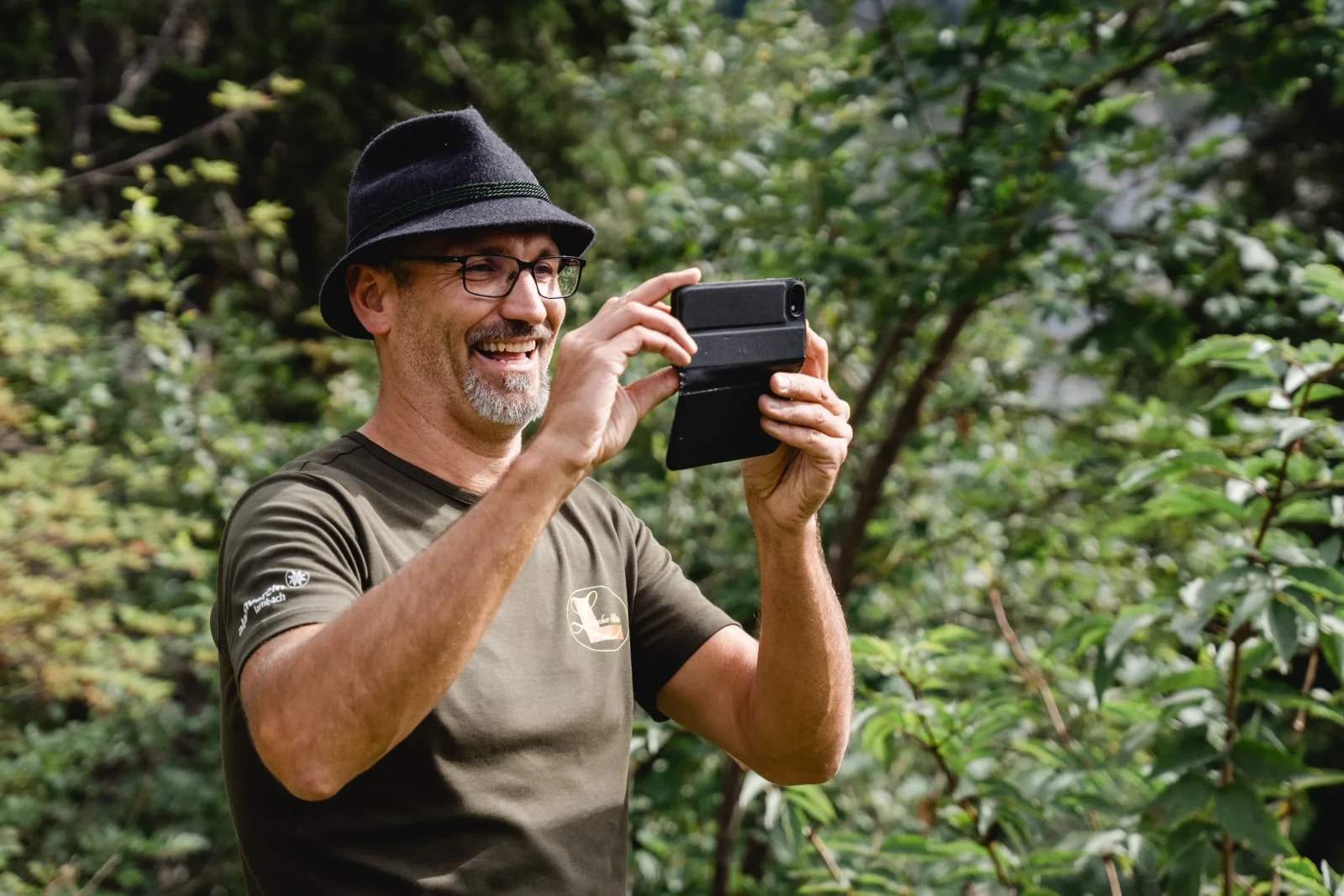 Lachender Mann mit Filzhut und Alpenverein-Shirt fotografiert mit dem Handy im Bergwald.