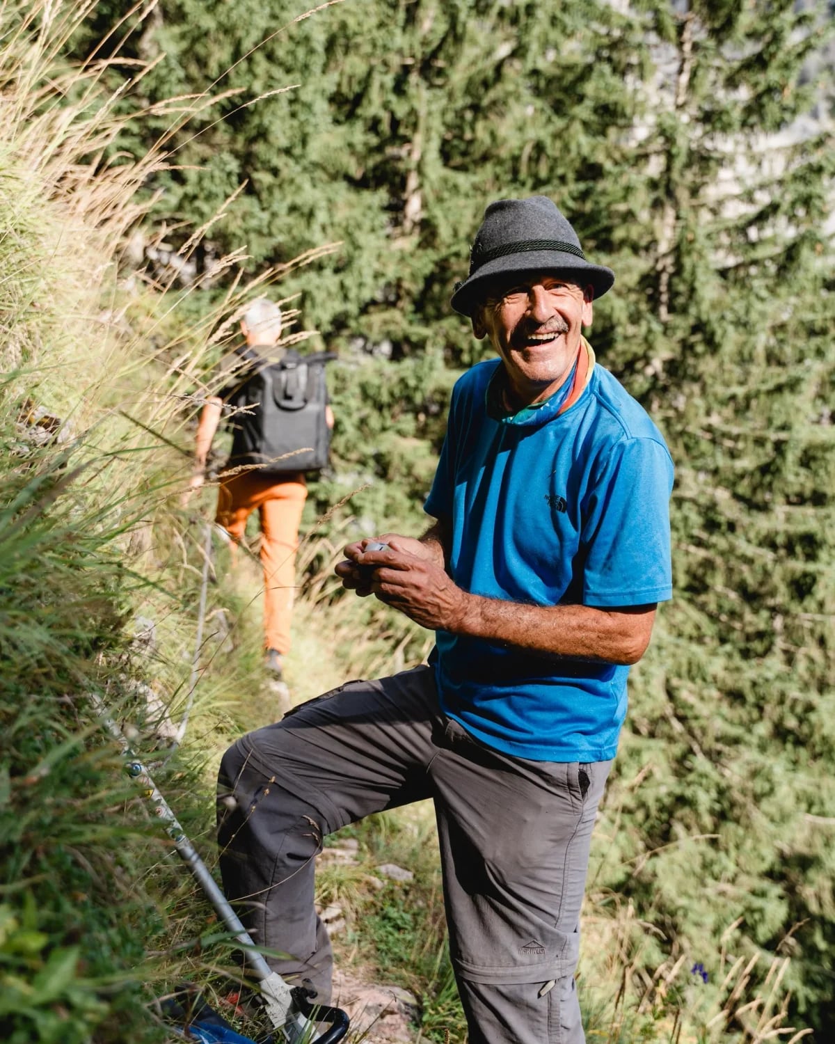 Lachender Mann mit Filzhut bei der Wegerhaltung am Bergpfad, im Hintergrund ein Wanderer und eine abgestellte Motorsense.