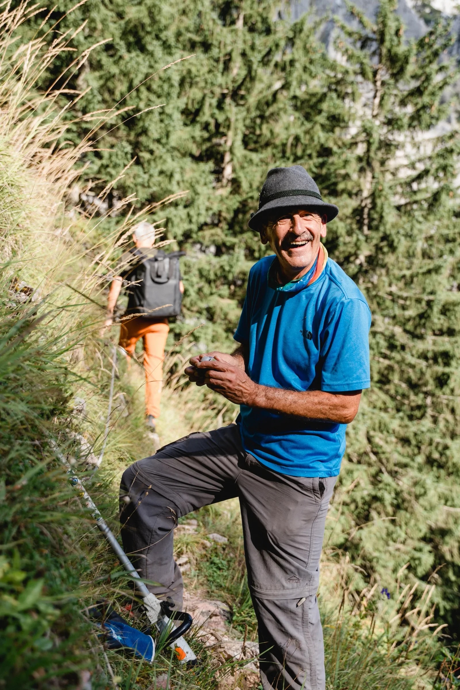 Lachender Mann mit Filzhut bei der Wegerhaltung am Bergpfad, im Hintergrund ein Wanderer und eine abgestellte Motorsense.