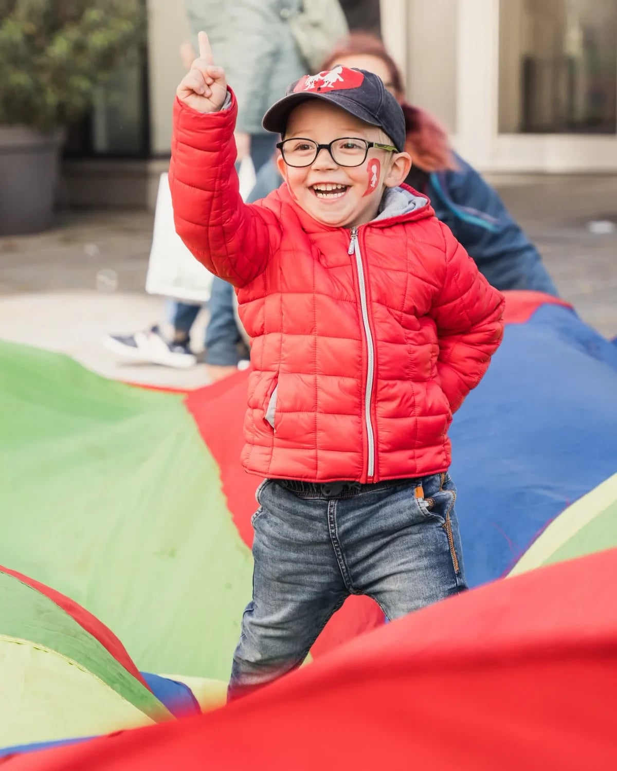 Lachender Bub mit Brille und roter Jacke reckt den Finger hoch, im Vordergrund ein buntes Schwungtuch beim Kinderfest.