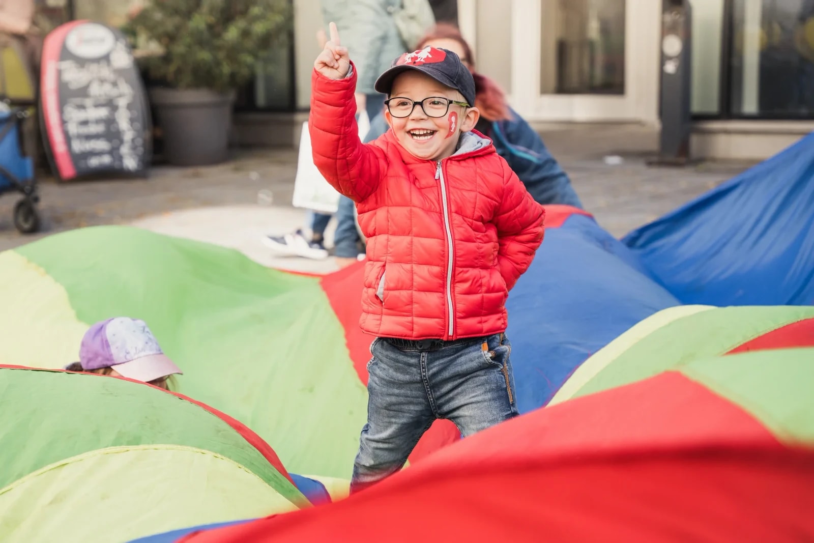 Lachender Bub mit Brille und roter Jacke reckt den Finger hoch, im Vordergrund ein buntes Schwungtuch beim Kinderfest.