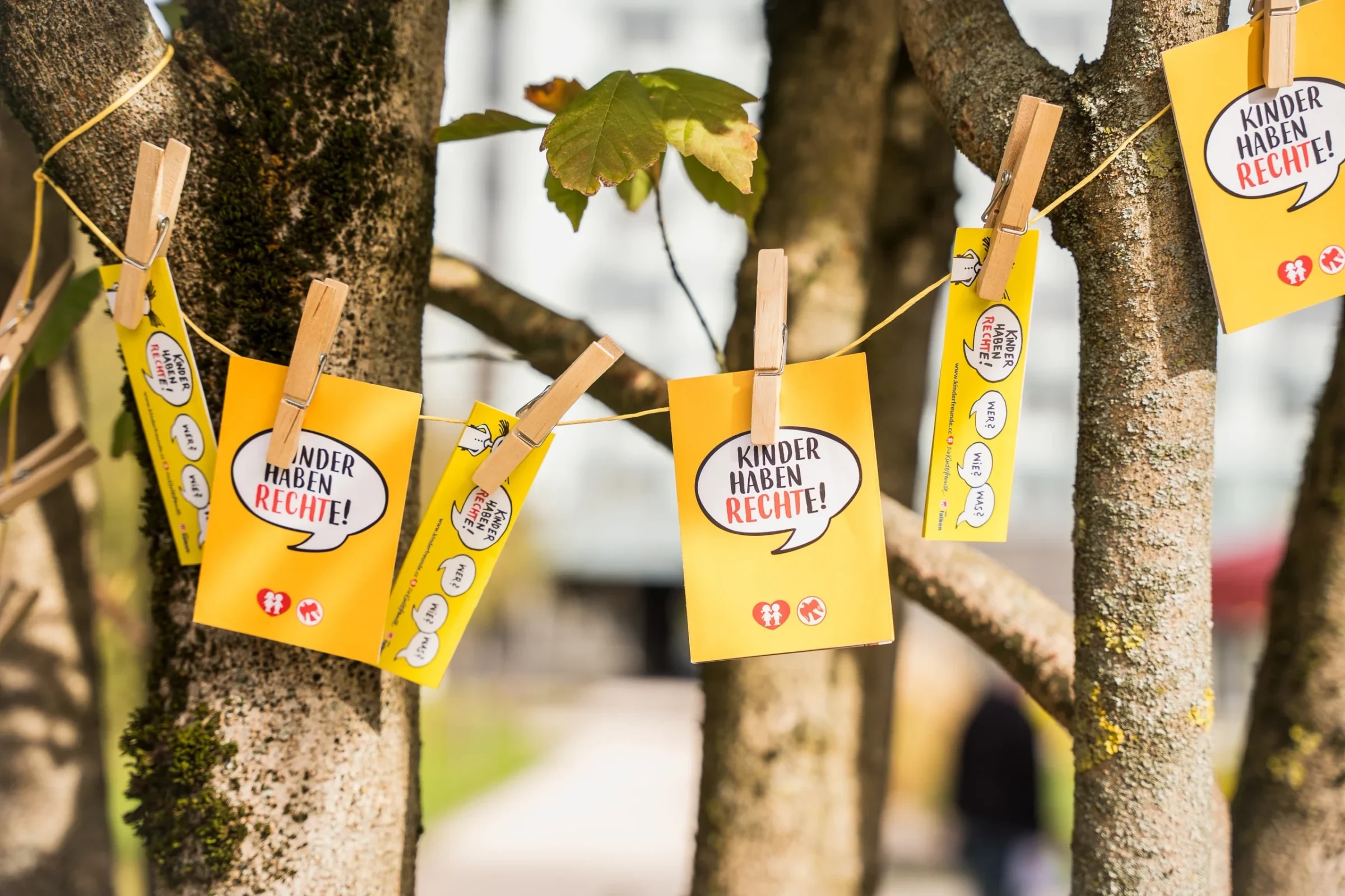 Gelbe Kärtchen mit der Aufschrift „Kinder haben Rechte!" hängen mit Wäscheklammern an einer Schnur im Baum.