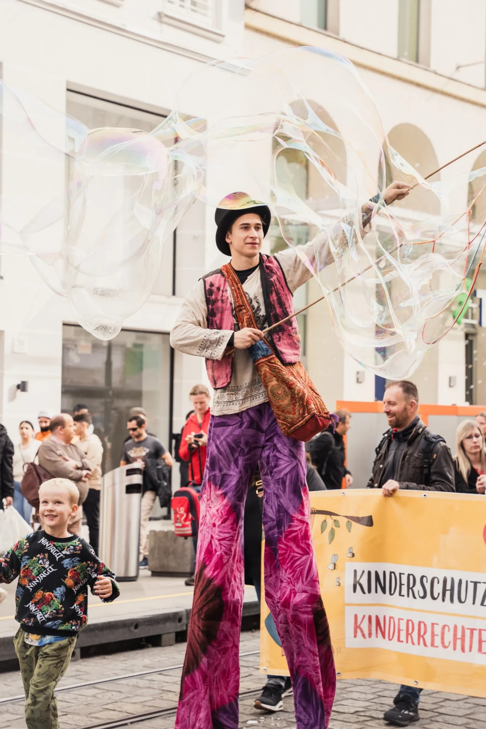 Stelzenläufer mit Riesenseifenblasen und ein lachender Bub vor dem Banner „Kinderschutz + Kinderrechte".