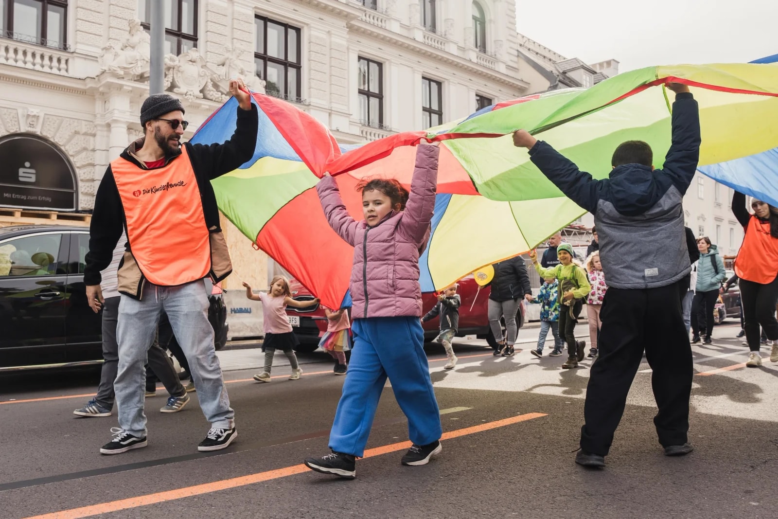 Kinder und ein Betreuer im Kinderfreunde-Leibchen tragen ein buntes Schwungtuch über die Straße.