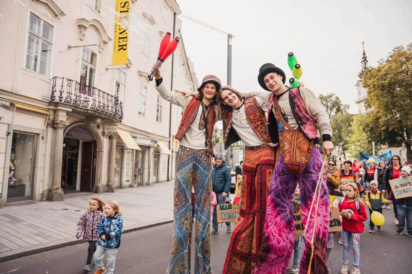 Drei Stelzenläufer in bunten Kostümen posieren vor dem Demozug der Kinderrechte-Demo.
