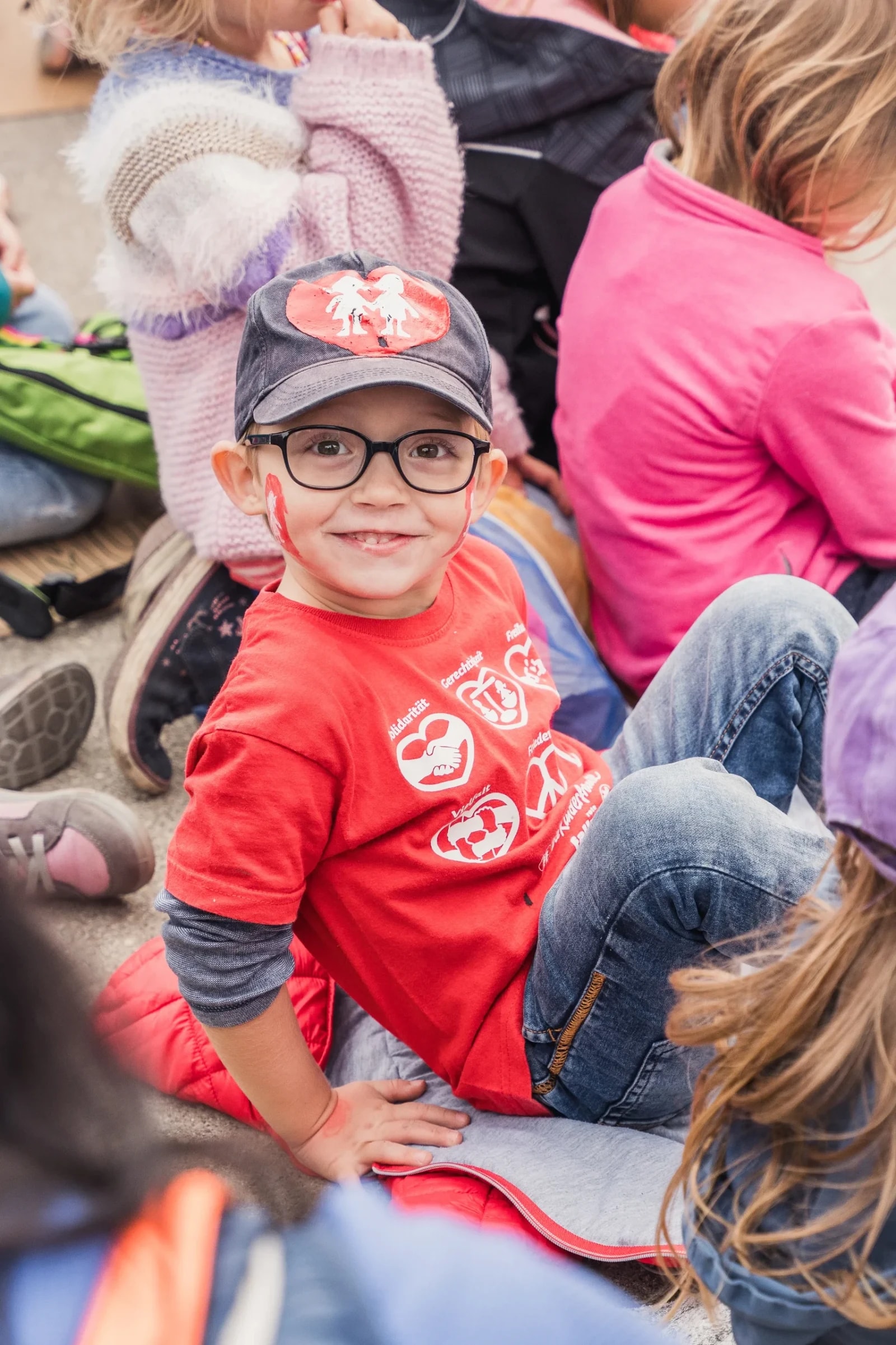 Kind im roten Kinderfreunde-Shirt mit Werten wie Solidarität und Gerechtigkeit sitzt in einer Gruppe am Boden.
