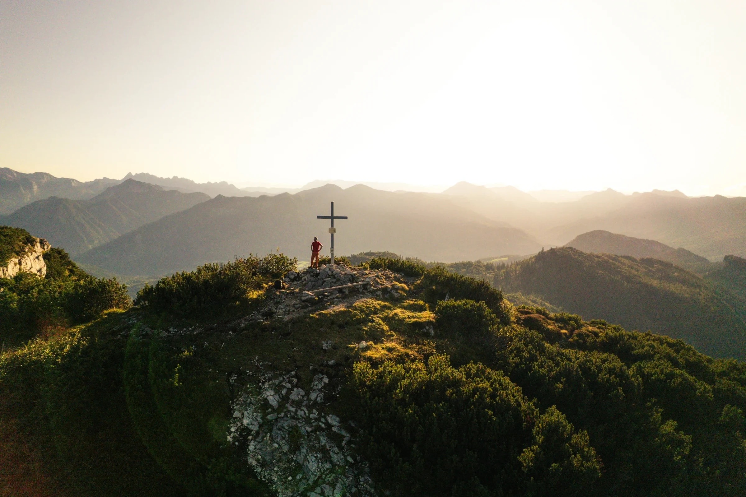Drohnenaufnahme: Person am Gipfelkreuz im Gegenlicht, dahinter Bergketten im Dunst der Abendsonne.