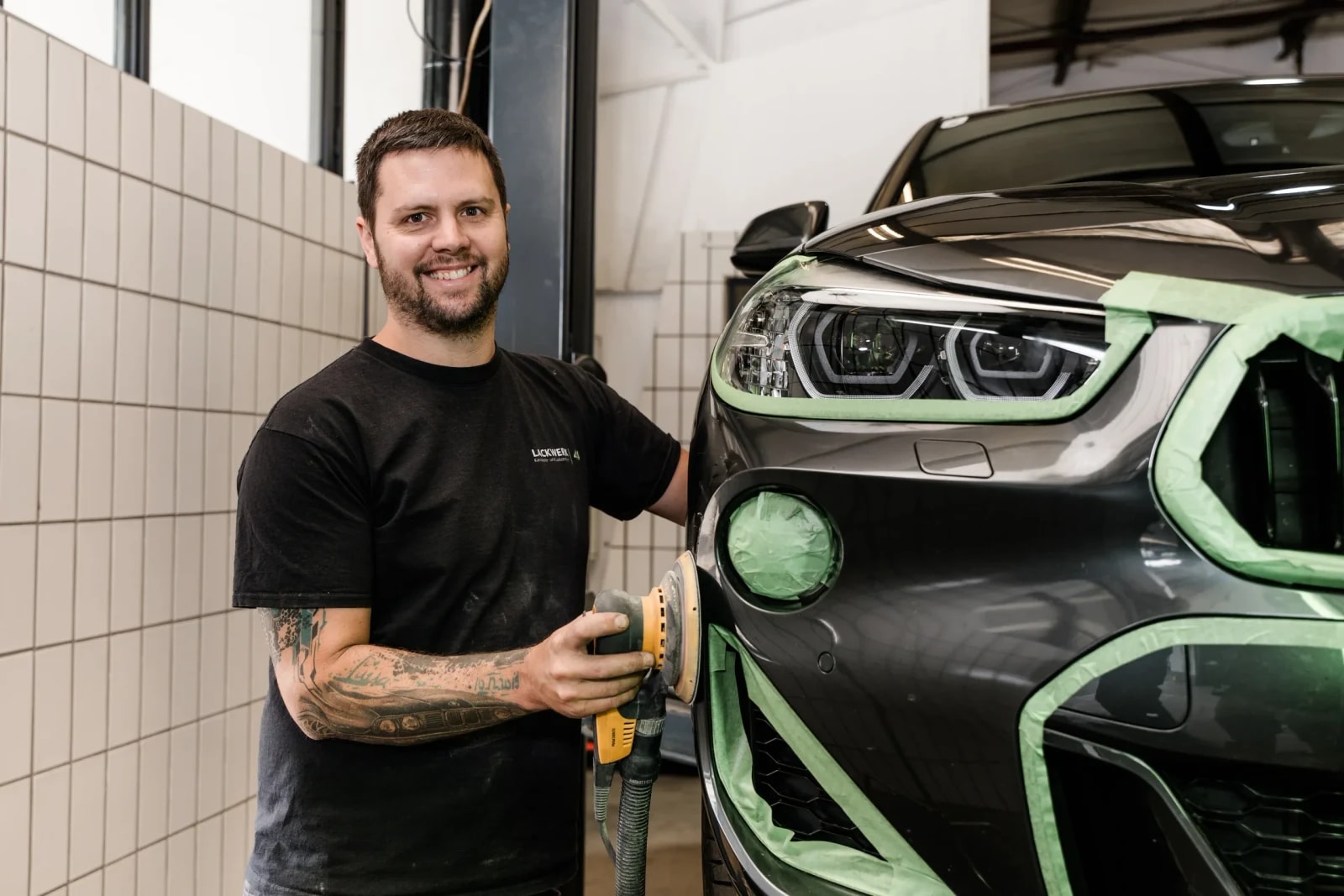 Karosserie- und Lacktechniker mit Exzenterschleifer neben einem abgeklebten Fahrzeug in der Werkstatt.