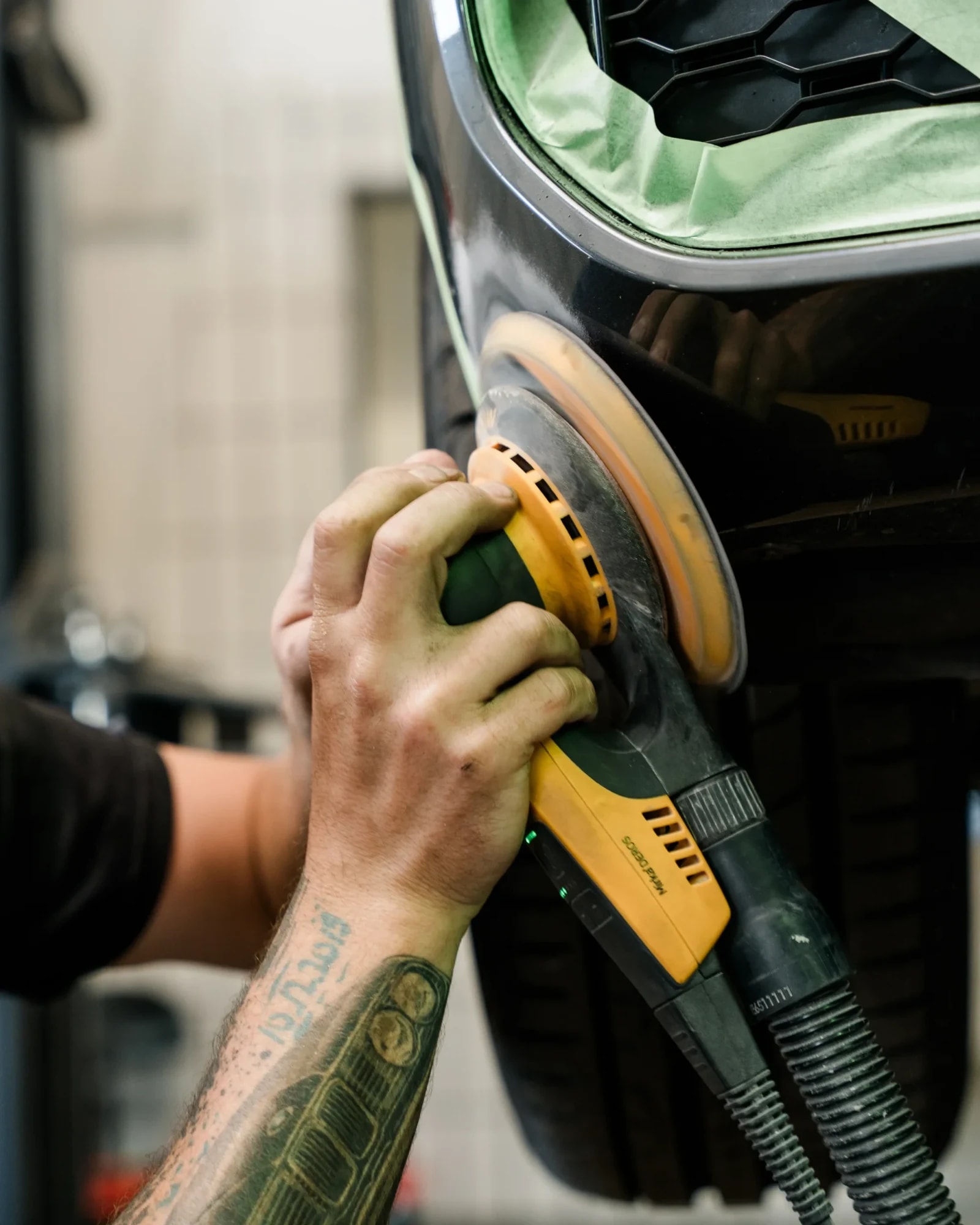 Detailaufnahme: Techniker schleift mit dem Exzenterschleifer einen Stoßfänger vor der Lackierung an.